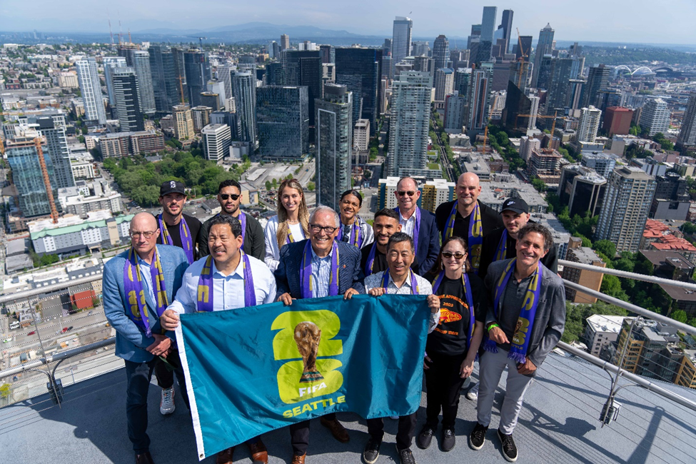 Gov. Jay Inslee and other leaders stand in a group above the Seattle skyline holding a World Cup flag.