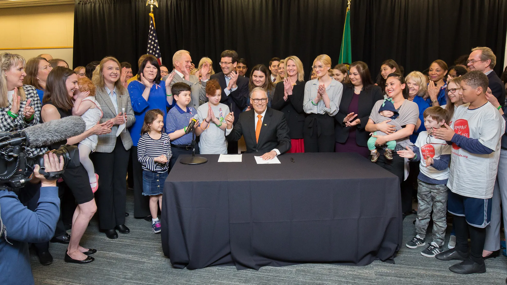 Several dozen people are clapping and smiling behind Gov. Inslee who is seated at a table signing a bill