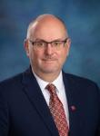 Drew Shirk, a man, wearing glasses, a blue suit, and patterned red tie