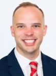 Portrait of Ryan Moran standing against a white backdrop, wearing a navy suit and red tie. 
