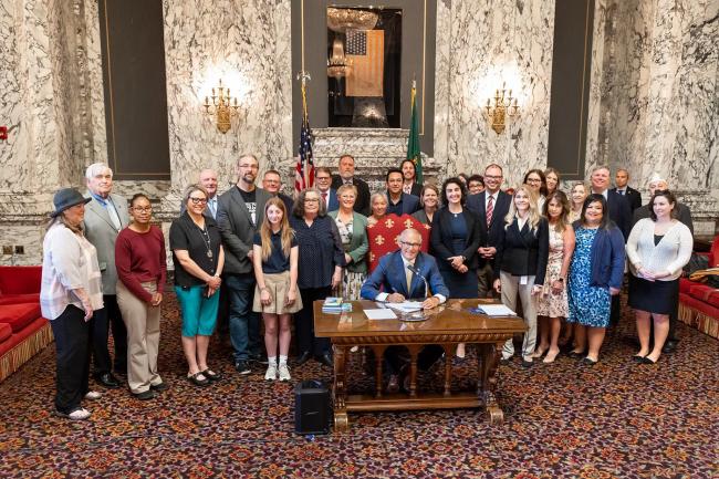 Gov. Jay Inslee signs a bill surrounded by advocates in an ornate state room.
