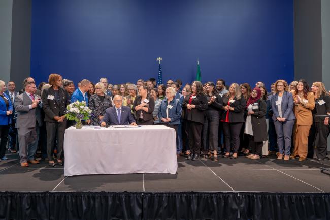 Gov. Inslee seated at a table with a large group of individuals standing behind him posing for a photo. 